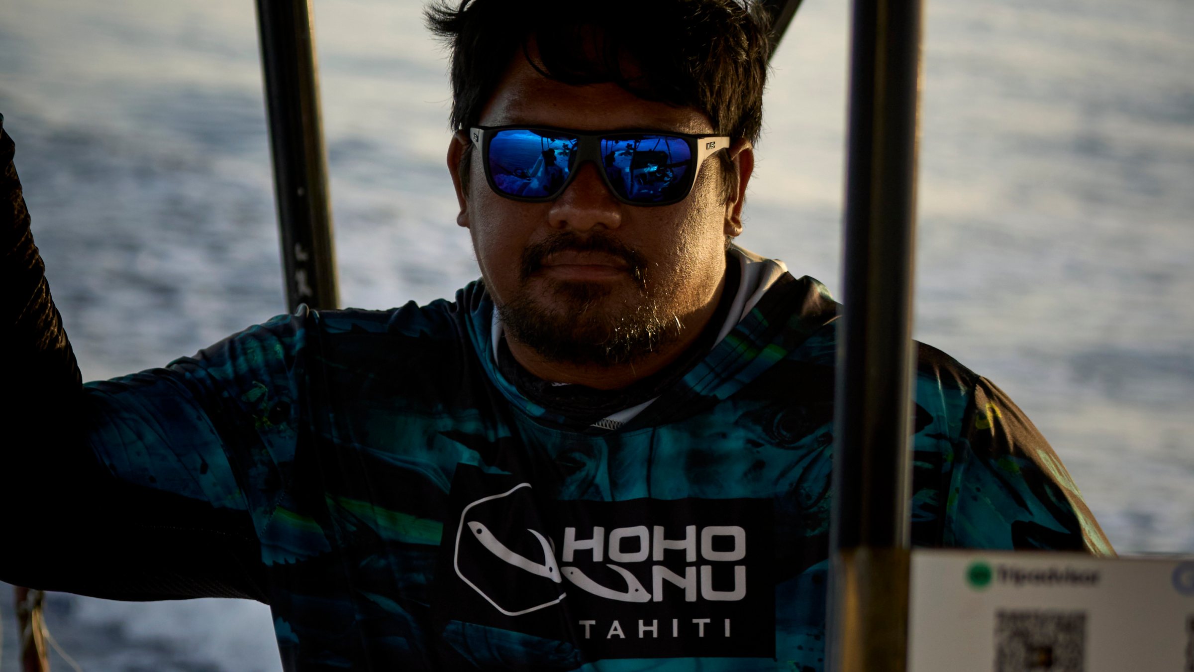 Man wearing sunglasses and printed shirt stands on a boat with the ocean in the background.