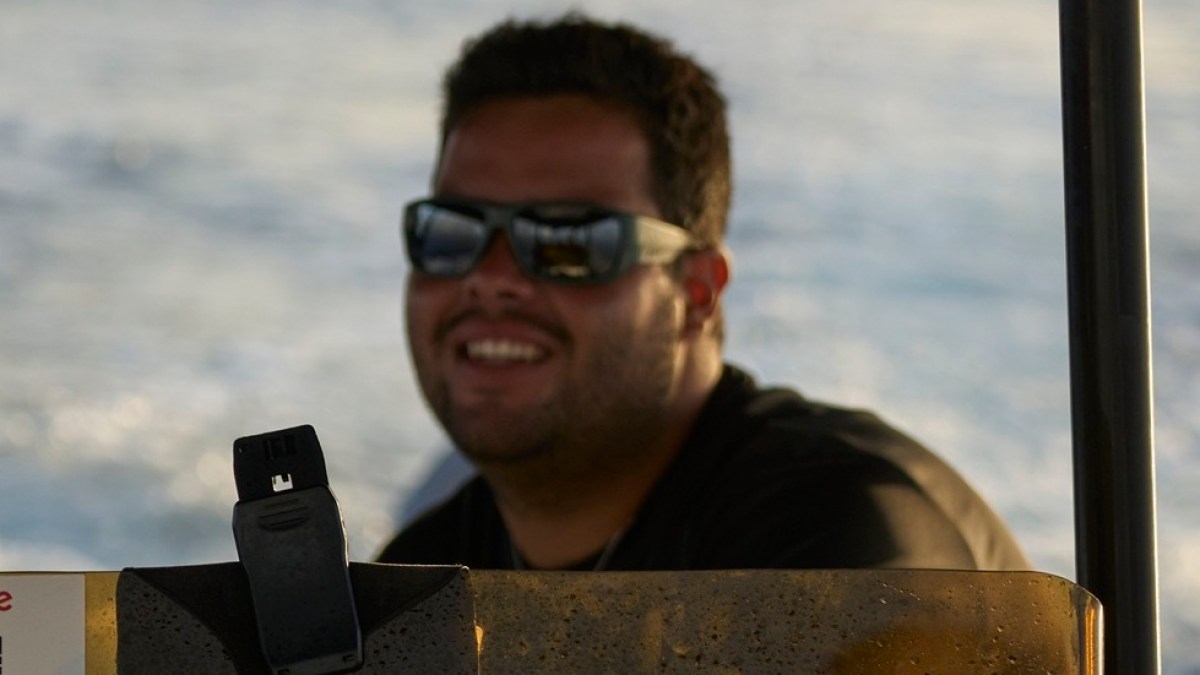 Man wearing sunglasses driving a boat, smiling, with blurred water in the background.