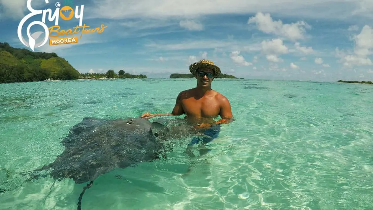 Man in sunglasses stands in clear ocean water with a stingray, wearing a straw hat and blue shorts.