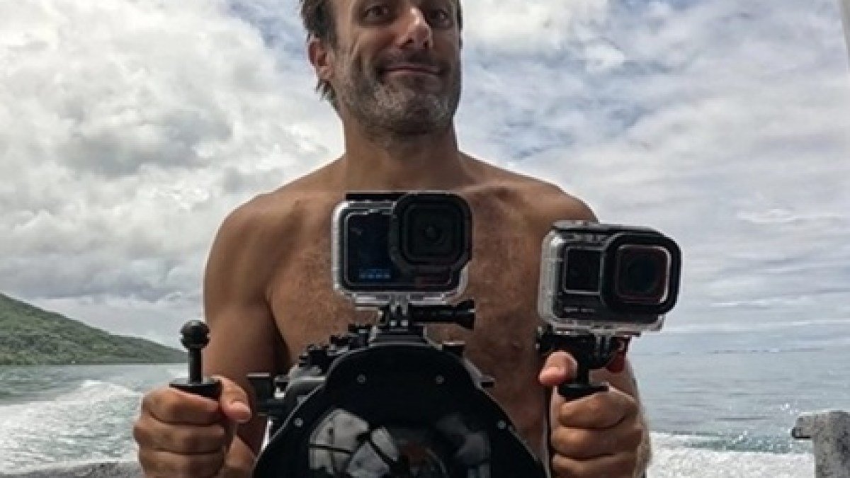 Shirtless man holding a camera rig with two action cameras on a boat, ocean in background.